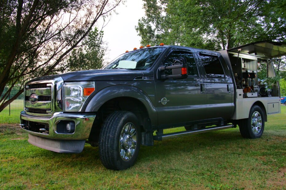 A side view of a dark grey 2014 custom Super Duty truck with open utility compartments, showcasing a mobile farrier workshop against an outdoor backdrop.