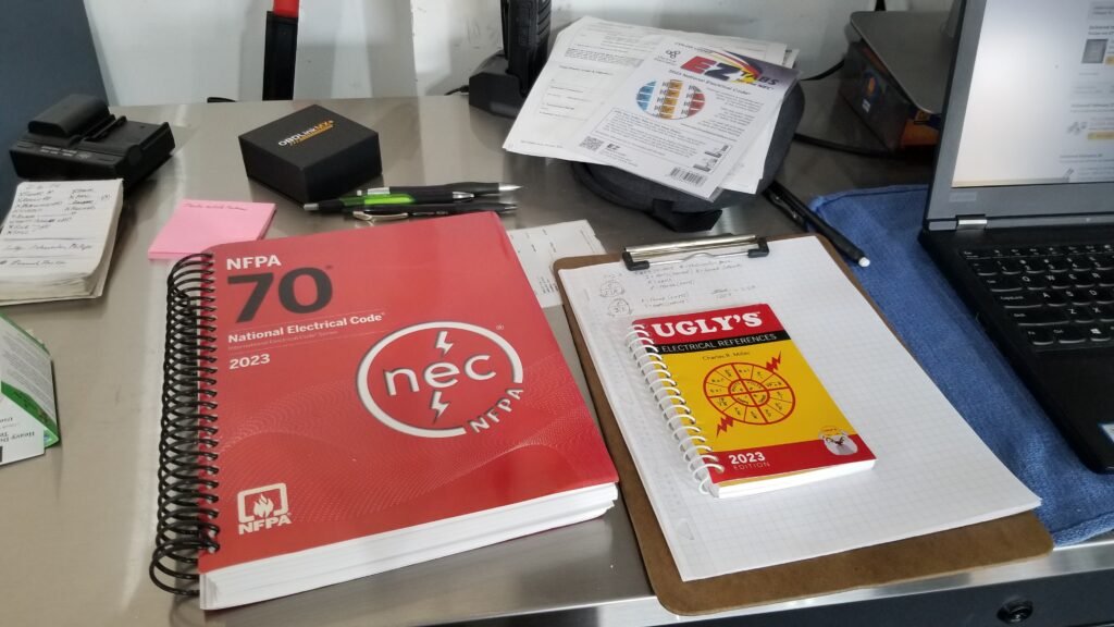 A professional electrician’s workspace featuring the 2023 National Electrical Code (NEC) book, Ugly’s Electrical References guide, a clipboard with notes, and various electrical study and testing materials.