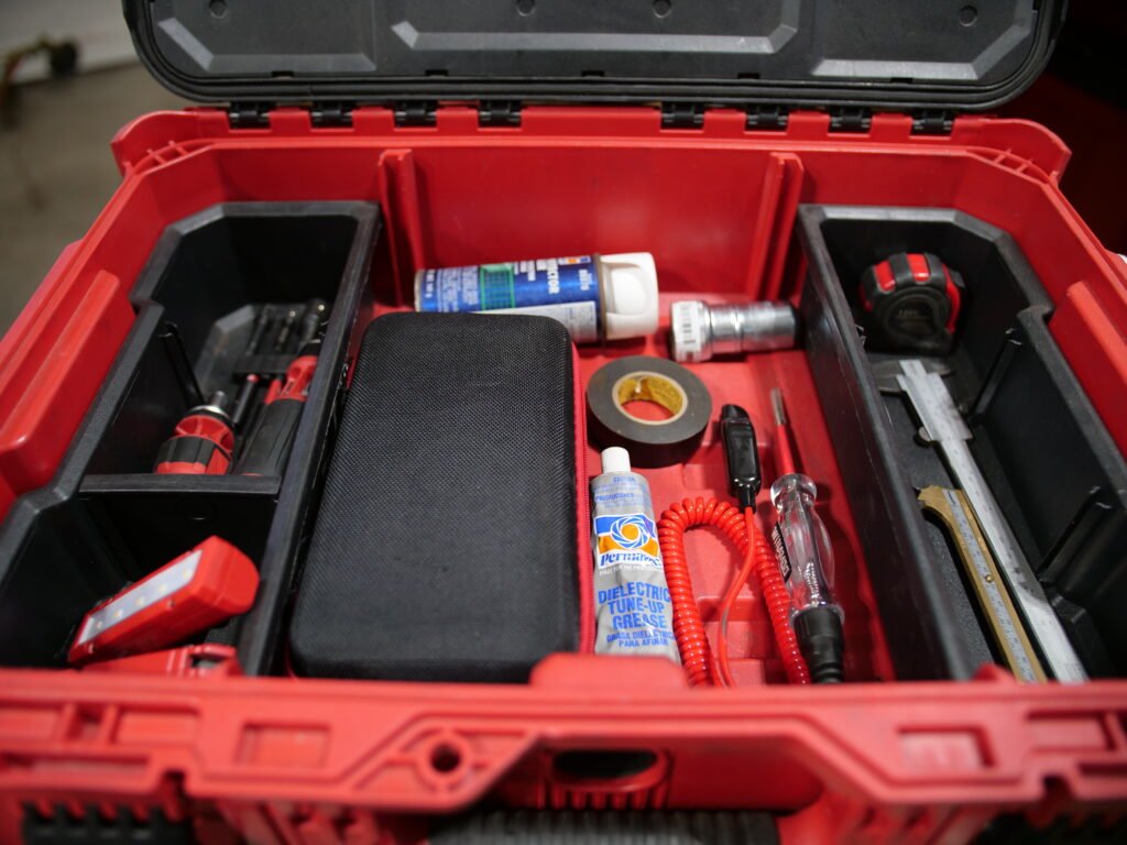Close-up of an open Milwaukee Packout compartment showing various tools and supplies, including screwdrivers, grease, tape, and a measuring tape.