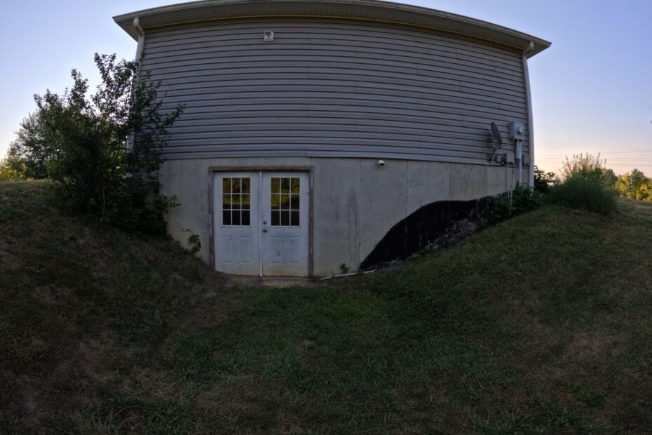 Basement entrance with double white doors set into a curved concrete foundation wall, surrounded by sloping ground and requiring retaining wall repair