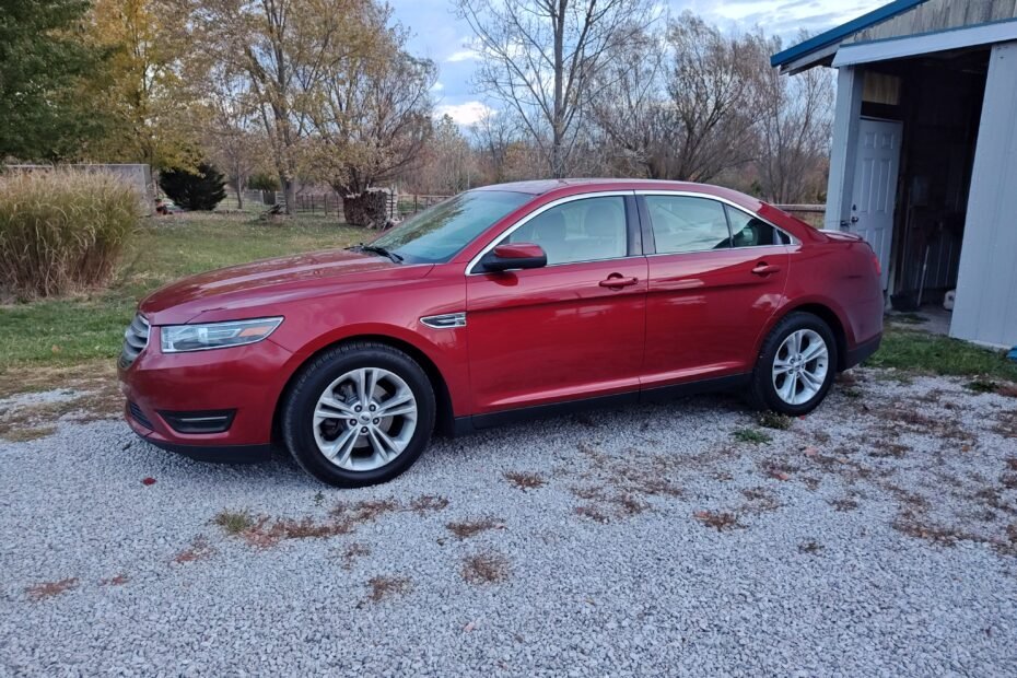 2015 Ford Taurus SEL in ruby red metallic parked on a gravel driveway, side profile view with autumn trees in the background