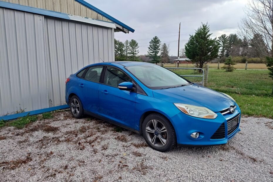 Blue 2012 Ford Focus parked on a gravel driveway beside a metal building, awaiting maintenance and transmission repairs.