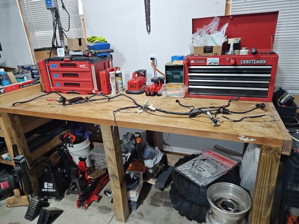 Disassembled wiring harness laid out on a wooden workbench in a well-equipped workshop, with tools and storage in the background.