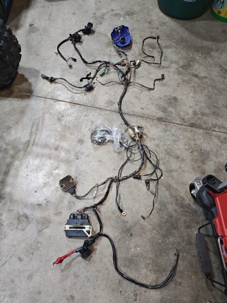 A disassembled 2005 Yamaha Raptor 660 wiring harness on a concrete floor, showing sensors, stator, and connectors, with visible wear and cuts in the wiring